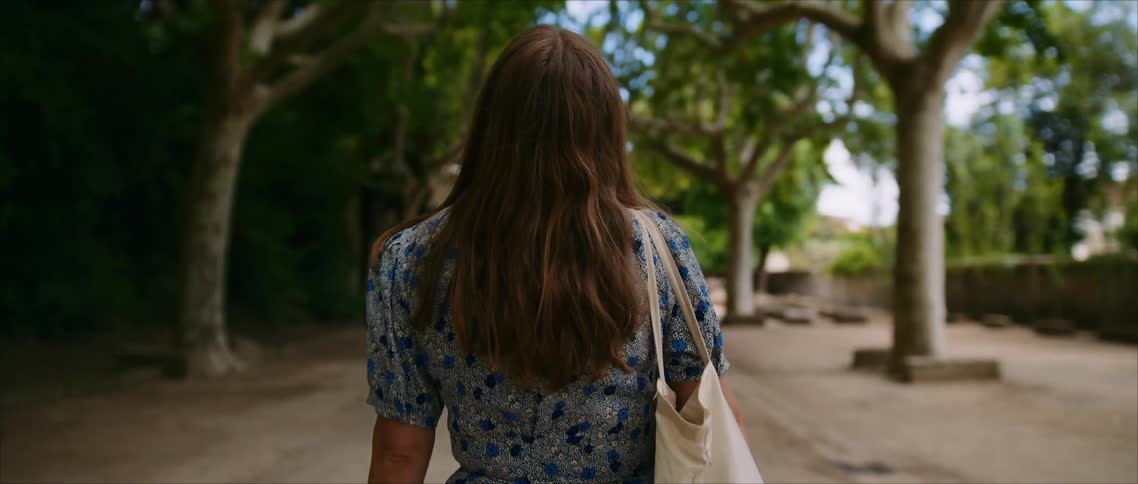A view from behind shows a woman with long brown hair wearing a blue patterned dress and carrying a white tote bag walking along a sunny path lined with trees and stone ruins
