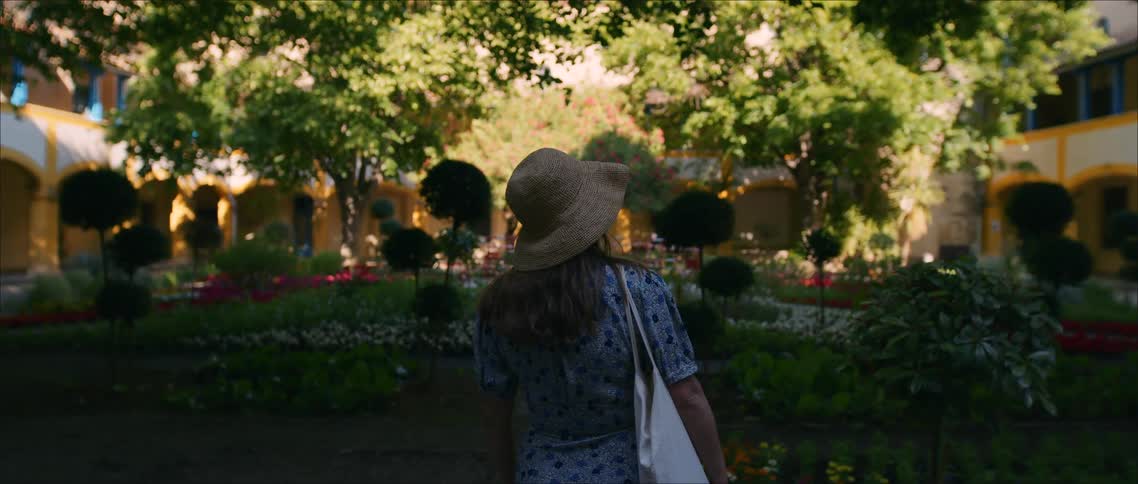 Woman wearing a hat walks through a flower garden surrounded by trees and arches
