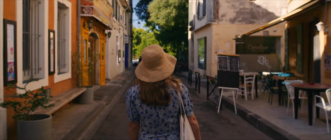Woman wearing a straw hat walks along a narrow street lined with cafs and colorful buildings