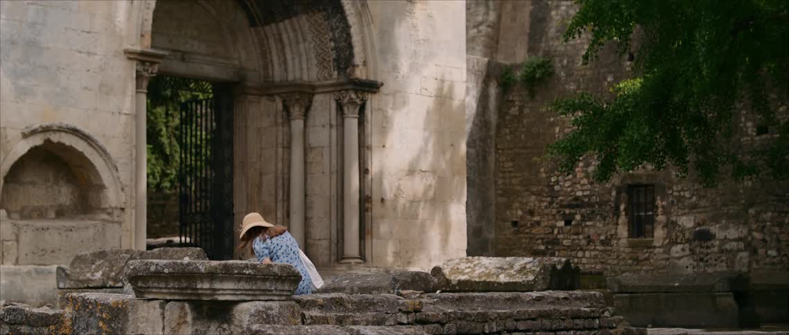 A woman wearing a straw hat and a blue patterned dress walks down stone steps and sits among the ancient stone ruins of a large abbey entrance on a sunny day