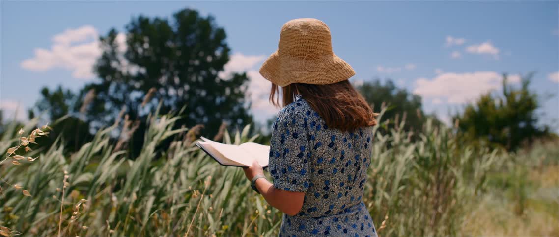 Person holds an open notebook in a tall grass field under a partly cloudy sky