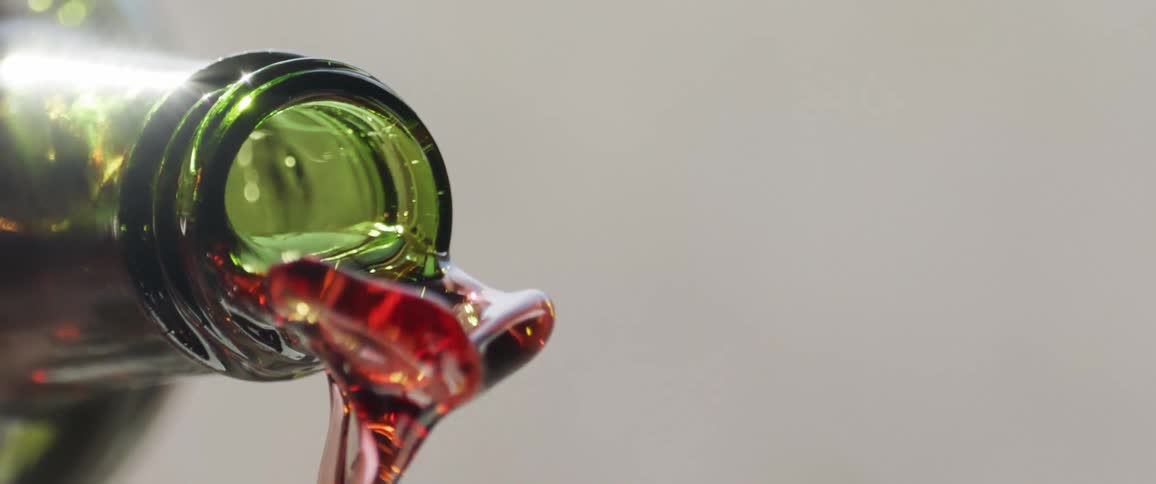 A close view shows a stream of dark red liquid pouring from the mouth of a dark green glass bottle against a solid light gray background