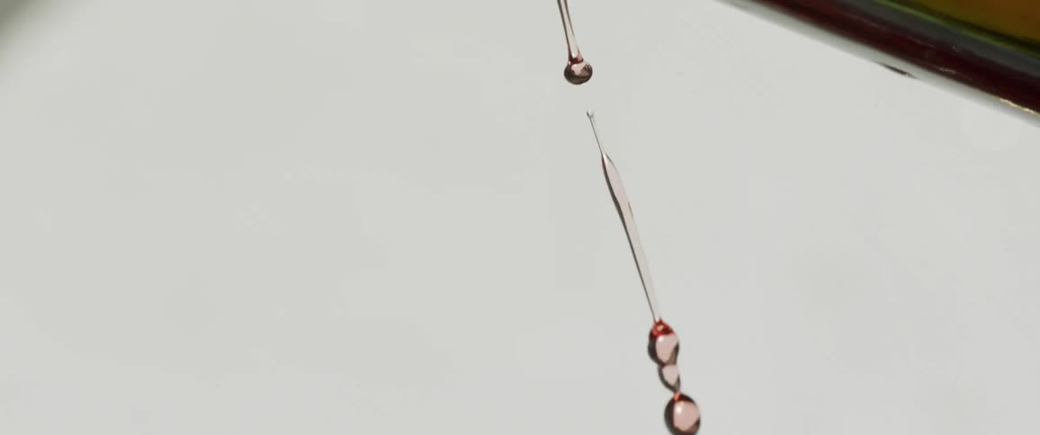 A close view shows red wine being poured from the neck of a dark green bottle thinning into a stream and then dropping off as individual droplets against a solid light gray background