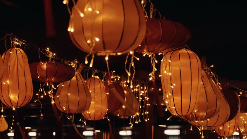 A view from below shows many glowing tear drop shaped orange paper lanterns hanging in the dark night intertwined with strands of small bright white fairy lights