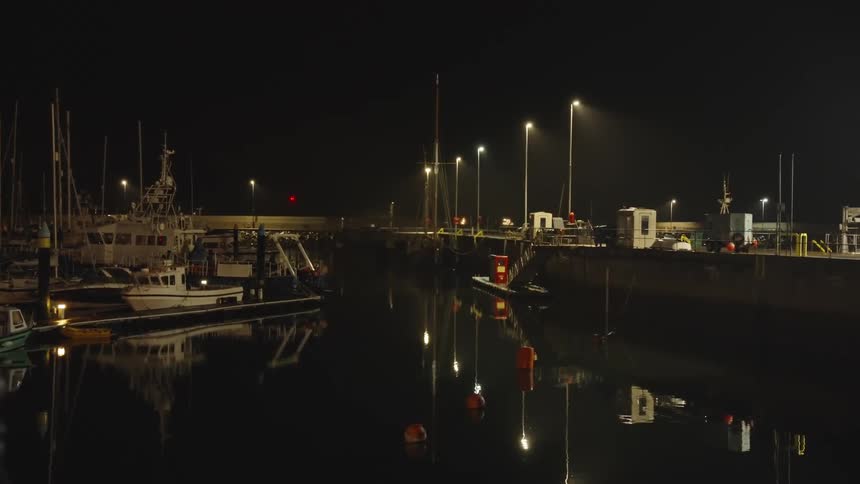 Docked boats reflect warm lights on calm waters in nighttime dock with vehicles in the background