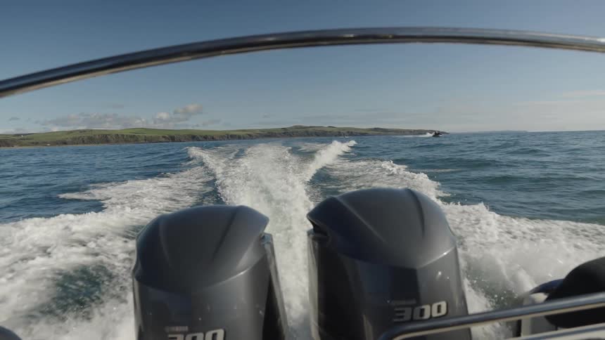 A view from the back of a boat shows two outboard motors running creating a white wake as the boat travels across the blue ocean toward a green coastal landmass under a clear sky