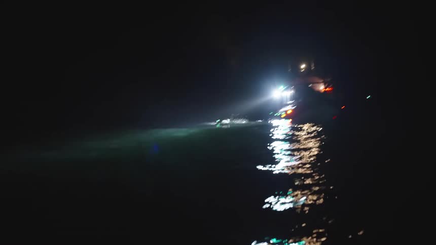 A small boat moving forward on dark water at night illuminating a swimmer with its bright forward facing spotlight that creates a long glistening reflection on the waters surface