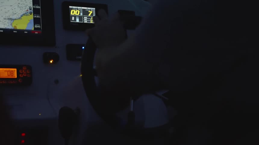 Person operates vessels wheel with nautical data displayed on screens during nighttime navigation