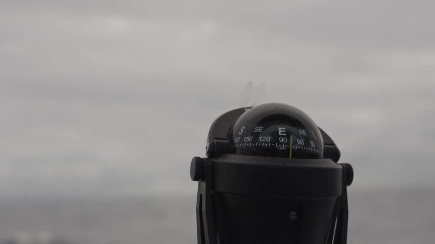 A closeup view of a black marine compass mounted on a surface shows the dial inside gently rotating against a bright featureless gray sky and water background