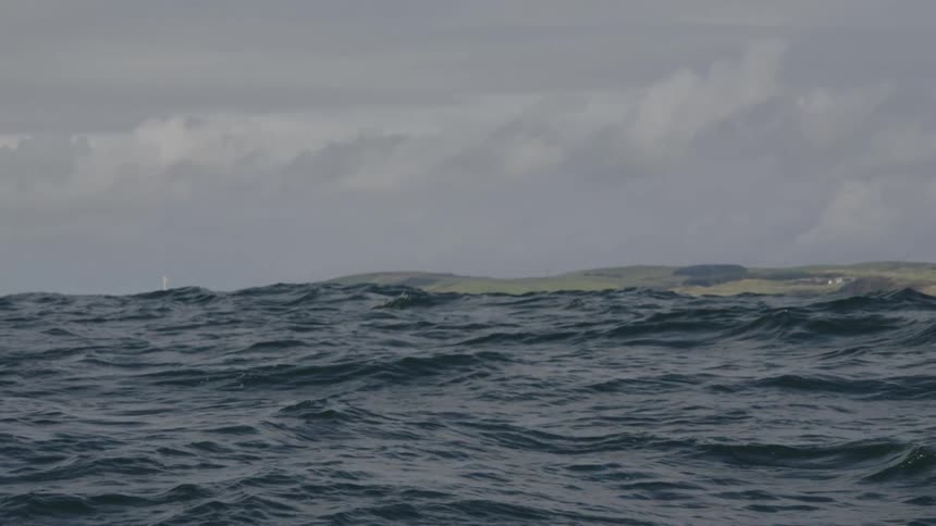 A choppy gray sea rolls beneath a cloudy sky with a low green coastline on the horizon