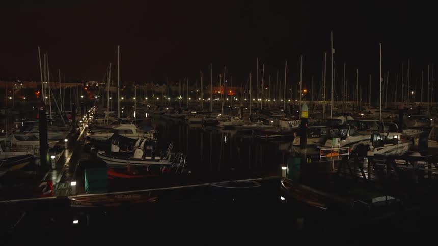 A boating dock sits quietly at night filled with boats and yachts gently bobbing in the water their silhouettes outlined by soft dock lights reflecting across the dark surface