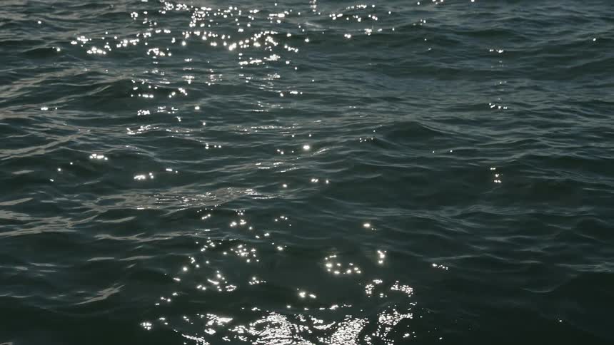 A full frame close view shows small waves of dark blue ocean water rippling rapidly as bright sunlight reflects off the surface creating many sparkling patches