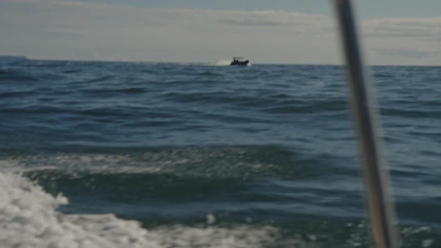 A view from a moving boat shows the churning water in the foreground and a distant motorboat traveling across the ocean waves under a cloudy sky framed by a metal boat railing