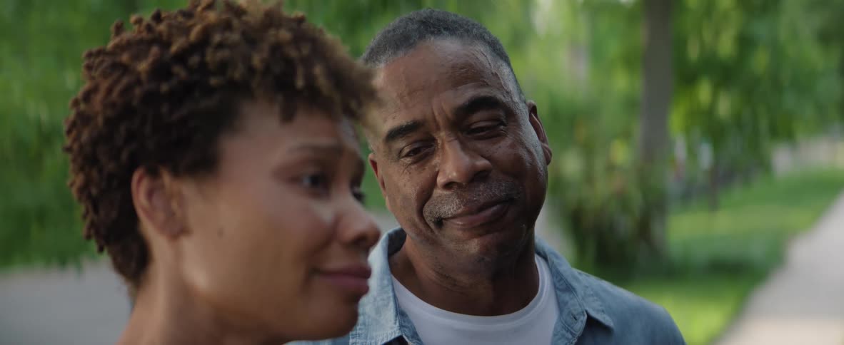 A close view of a smiling older man with gray hair and a younger woman with curly brown hair looking off to the side while standing outdoors in a sunny park