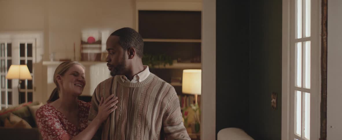 A black family shares a warm moment in their living room the mother walks over to the father gently resting her head on his shoulder as he picks up a sweatshirt just then their daughter playfully snatches the shirt from his hands prompting a smile