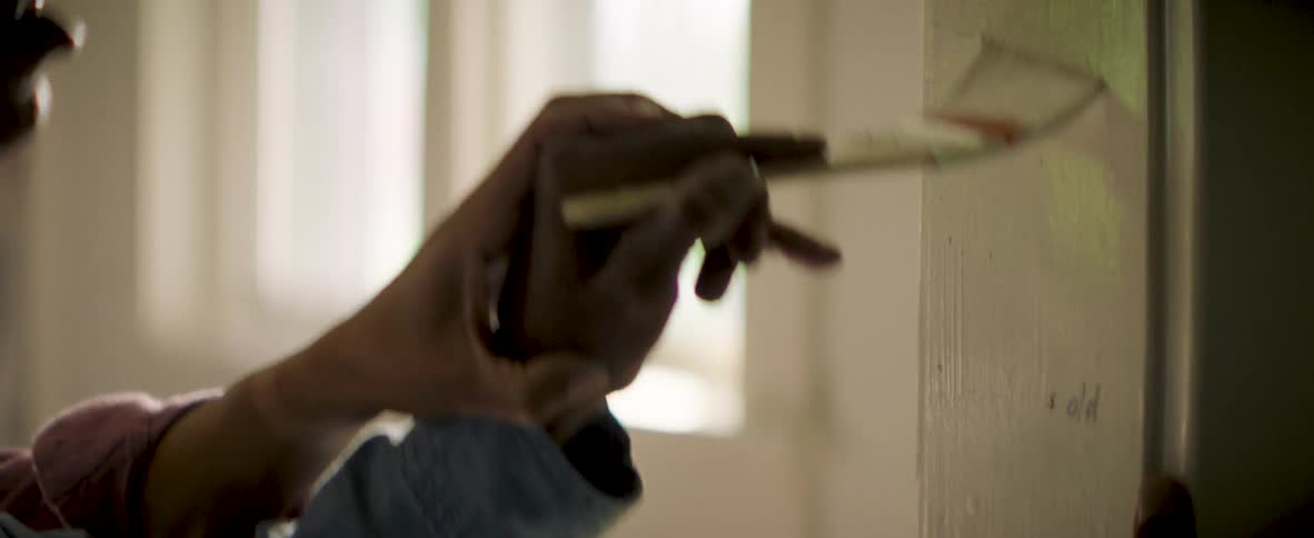 A hand on a hand holds a paintbrush to a doorframe marked with height lines and ages in a softly lit room