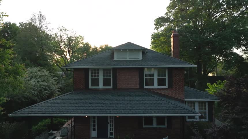A two story brick house with a broad gray shingle roof is framed by tall trees with sunlight peeking over the roof peak and a red brick chimney on the right