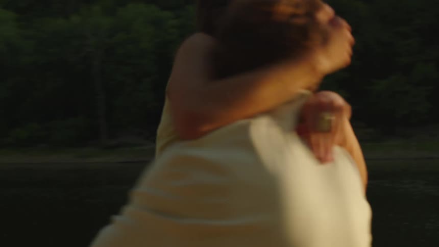 Man picks up a woman and spins her around while standing on the bank across the river from the woods