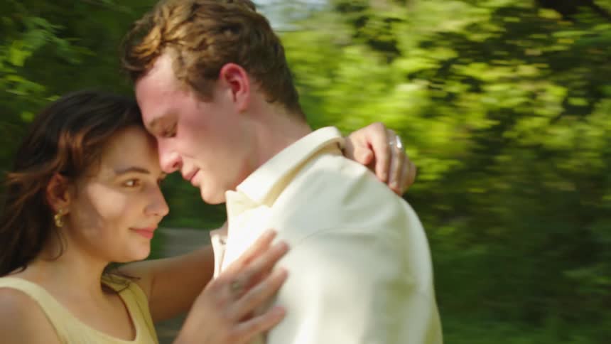 A young couple in light clothing embrace with their foreheads touching and tender expressions as they sway together outdoors against sunlit greenery