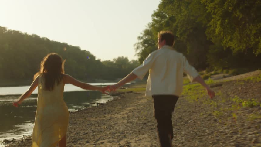 A couple runs handinhand along a rocky riverside at golden hour