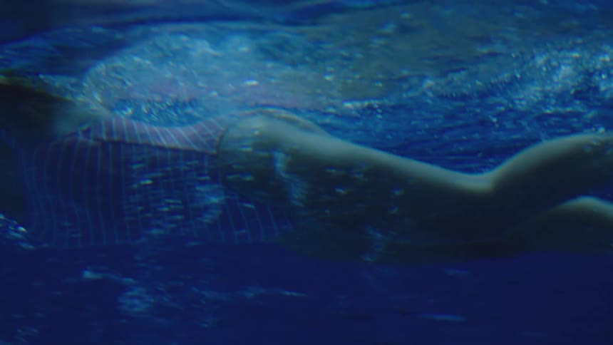 The video is an underwater shot of a persons body swimming in a pool or deep body of water the lighting is subdued giving the water a deep uniform blue color and highlighting the silhouette of the swimmer