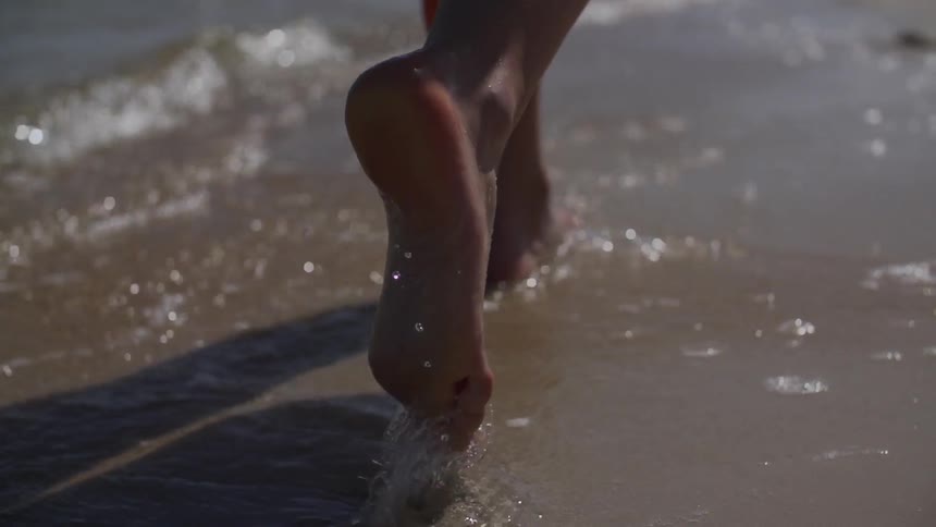 Close up shot of someone walking at the edge of the water on a beach as waves gently was on shore