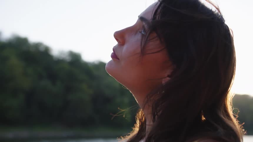A woman in profile looks toward the low sun by the water backlit with a calm contemplative expression