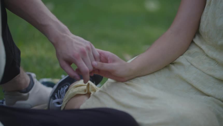 A young couple sits hand in hand on a sunlit grassland at midday sharing a quiet romantic moment surrounded by nature