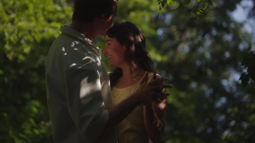 A man kisses a womans hand beneath leafy shade their faces close with a tender affectionate mood