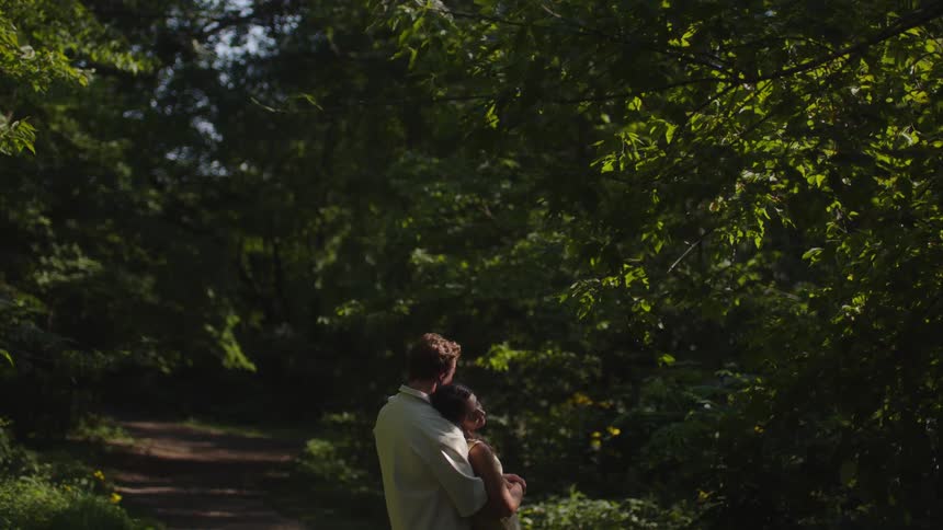 A man is holding a female in his arms as they both stand and sway in a wooded area on a path