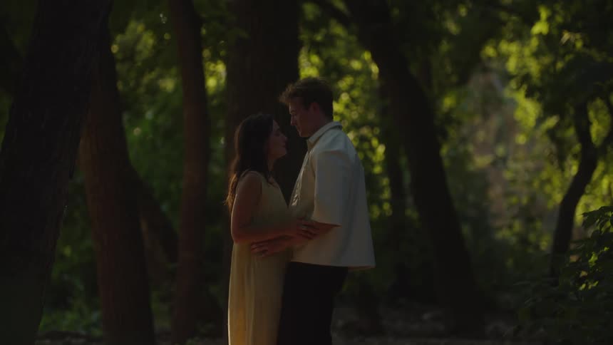 A young couple embraces beneath a tree in the forest at midday sunlight filtering through the leaves and casting warm patterns across the ground as they share a quiet moment together