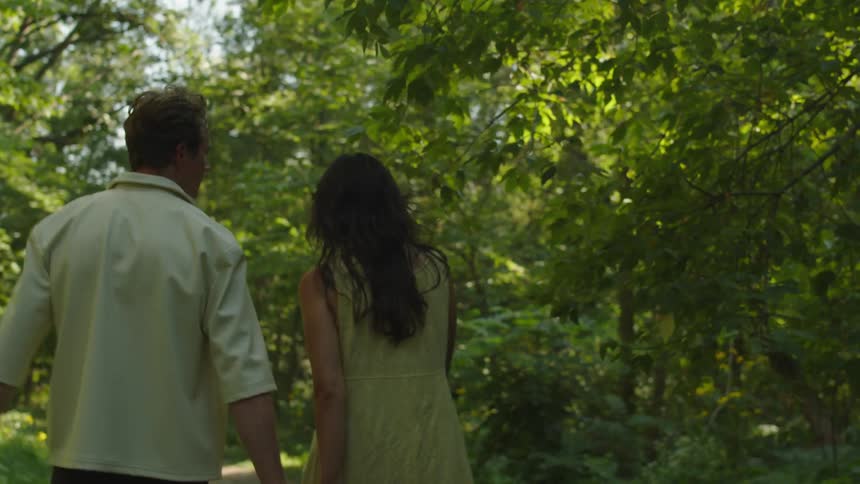 Man and woman seen from behind walk together in green area with sunlight filtering through trees