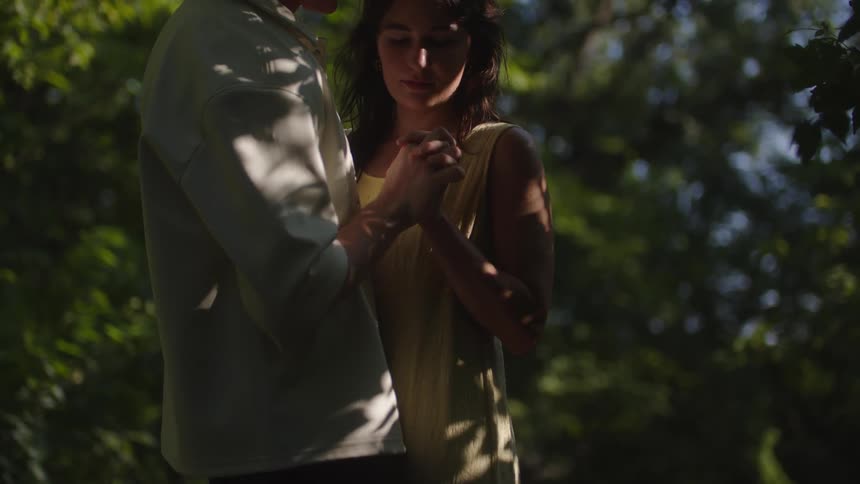 A man in a light colored shirt and a woman in a yellow dress embrace in a forest clearing as she looks up and smiles at him while dappled sunlight shines through the green leaves