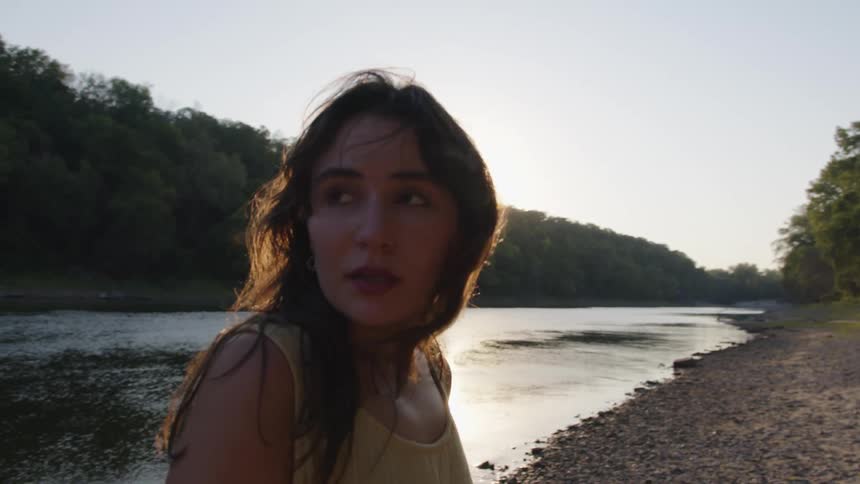 Woman walks by a riverside at sunset glancing back with a gentle smile