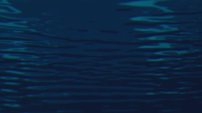 An underwater view looks up at the surface of the water as abstract light ripples reflect off the ceiling of a dark blue pool or ocean environment