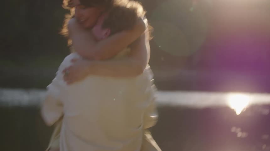 A man in a white shirt carries and spins a woman in a yellow dress while embracing her with bright sunlight flaring over a dark lake and rocky shore in the background