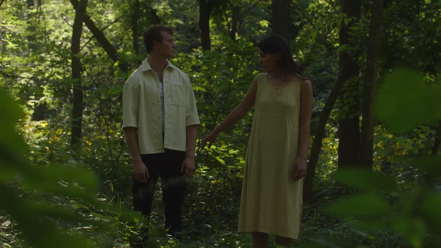 The video shows a man and a woman standing in a deeply wooded forest clearing facing each other the woman is wearing a light yellow dress while the man wears a lightcolored buttonup shirt and dark pants