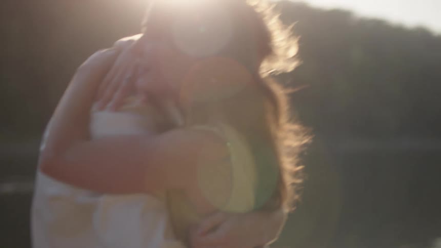A young couple enjoys a playful outdoor moment on a riverbank at sunset hugging each other warmly as golden light reflects off the water their faces glow with smiles surrounded by the peaceful beauty of the fading day