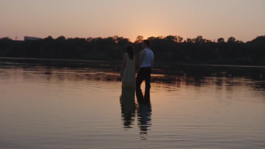 A couple stands in ankle deep water holding hands watching the sunset