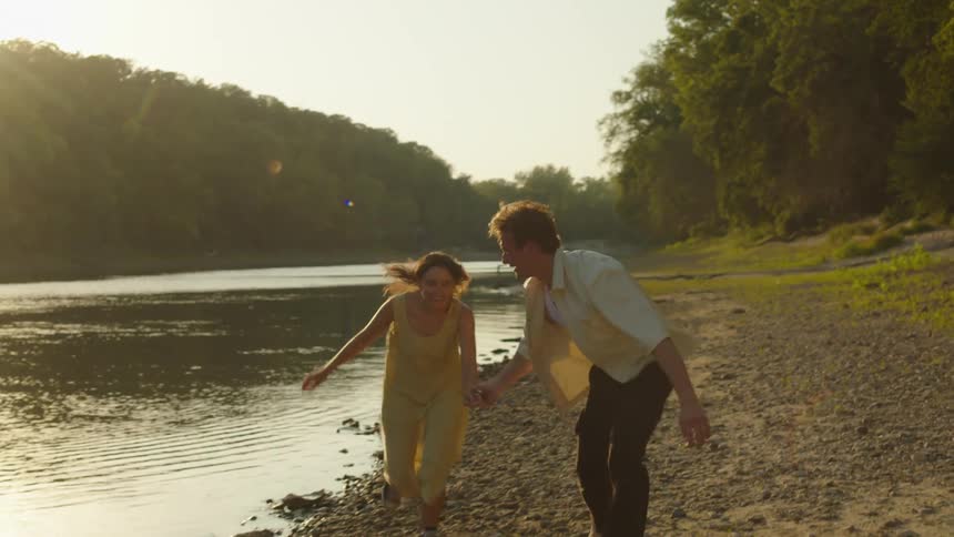 A young couple runs hand in hand along the riverbank in the woods at sunset their silhouettes framed by golden light filtering through the trees as the water glows beside them