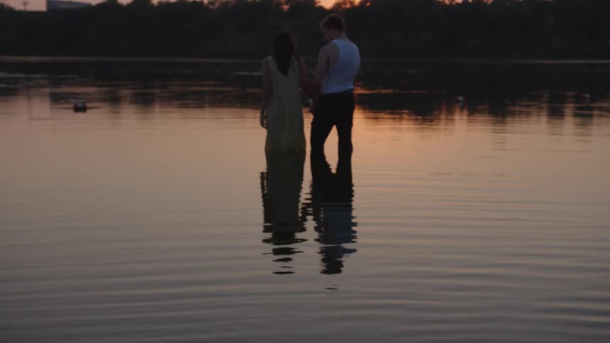 A female and male adult couple are standing foot deep in a lake nears woods at dusk while holding hands