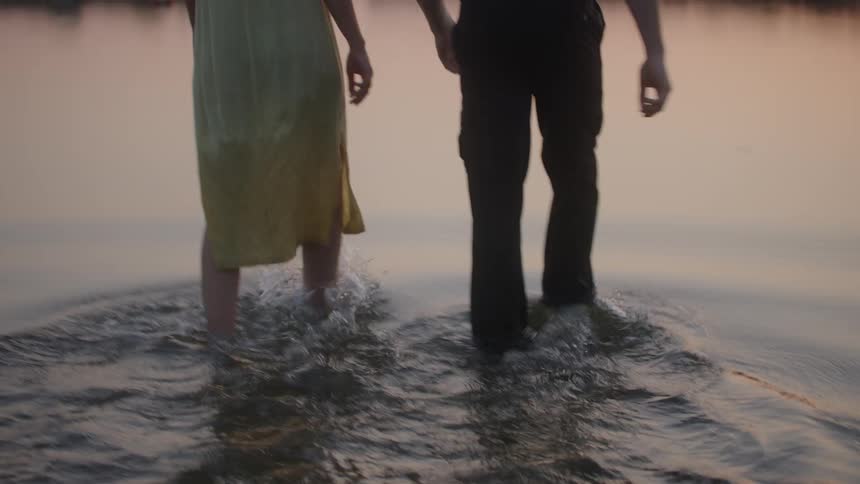 The legs and bare feet of a man in dark pants and a woman in a long yellow dress are seen from behind as they walk through shallow water on a beach at sunset