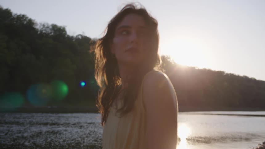 A woman in a pale yellow dress stands by a lake or river with sunlight directly behind her creating a bright lens flare and a silhouetted view of her and the surrounding forested hills