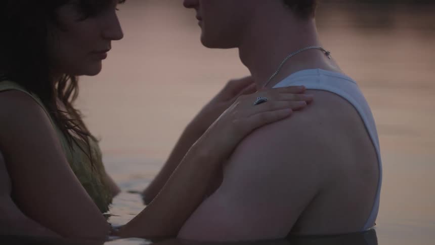 Man and woman stroke each others skin and nuzzle together while standing in water tinted red by the sunlight