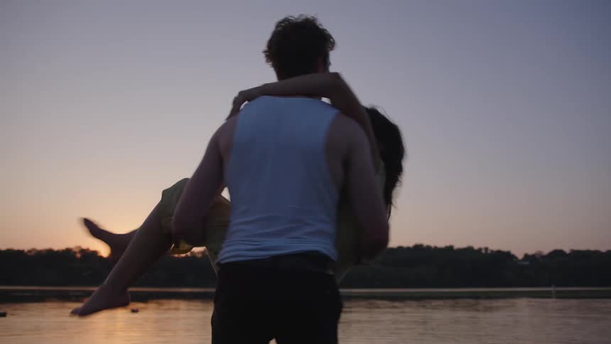 Man carries woman in his arms as they laugh in the water at sunset