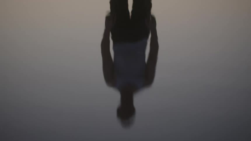 The upside down silhouette of a person is perfectly reflected in a body of still water creating an abstract symmetrical image against a light gray sky