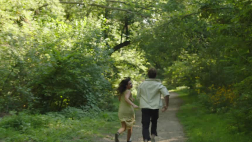 A handheld shot follows a joyful young couple from behind as they run hand in hand down a path through a lush forest