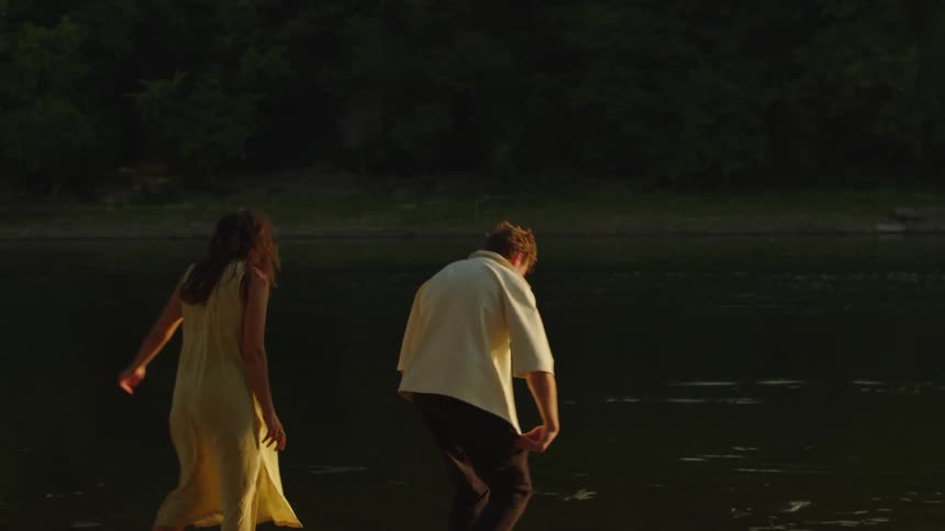 A man in a light shirt and dark pants stands on a rocky shore with a woman in a yellow dress as they both turn toward the dark water throwing rocks and enjoying the golden sunset light