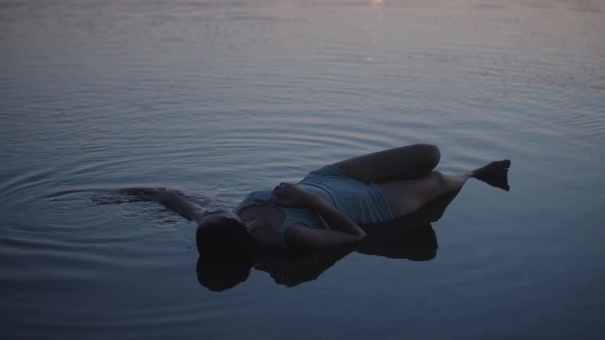 A woman floats on her back in dark still water with ripples and reflection at dusk