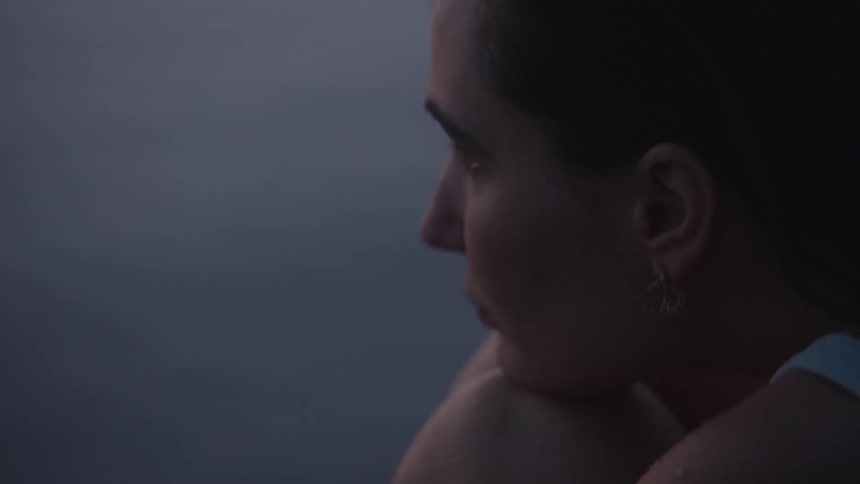 A closeup shot of a womans face in profile shows her resting her head on her bent arm or knee with a solemn expression against a body of water